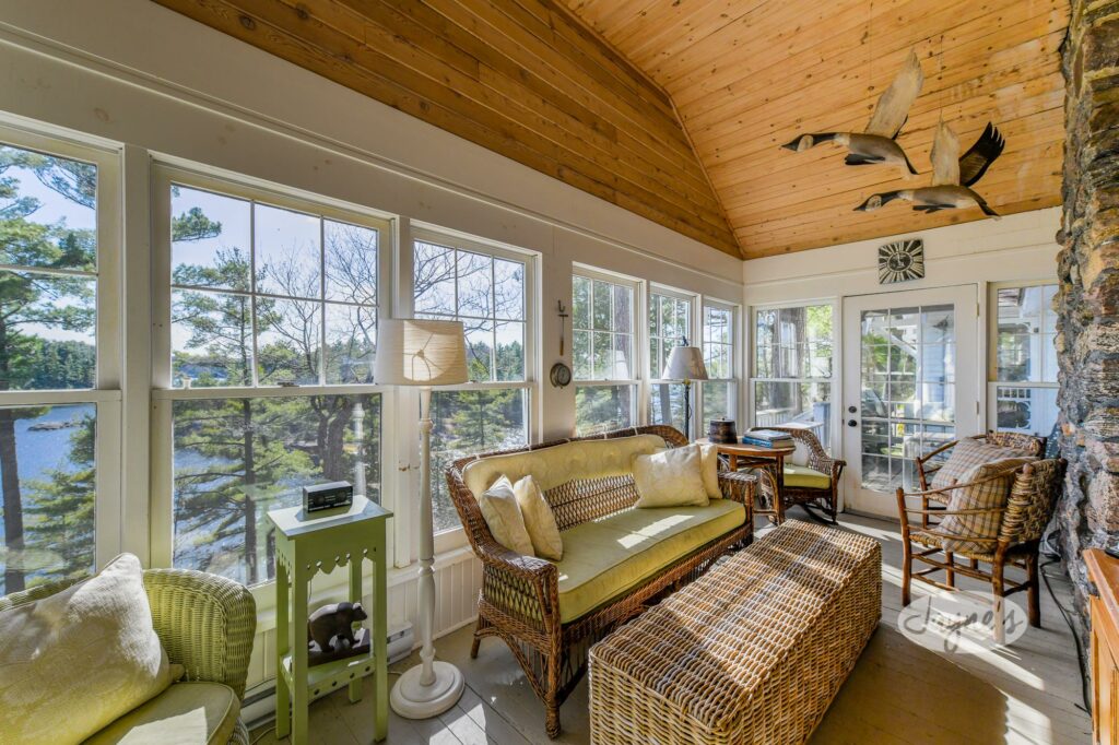 Bright sunroom with panoramic views of the lake, featuring cozy wicker furniture and natural wood accents - Jayne's Luxurious Rentals.