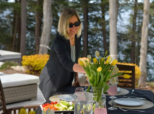 Elegant outdoor dining setup by Jayne's Luxury Rentals with a smiling host arranging yellow tulips on a lakeside table, enhancing the gourmet experience.